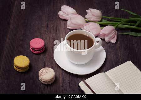 Tazza di tè, macaron, tulipani rosa e taccuino su sfondo di legno. Foto Stock