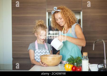 Famiglia, cucina, cottura e concetto di gente - madre felice con archetto sui suoi capelli crespi con figlia piccola con ciotola di legno che versa l'acqua e mak Foto Stock