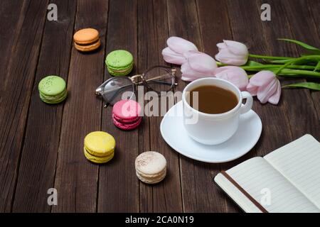 Tazza di tè, macaron, bicchieri, tulipani rosa e taccuino su sfondo di legno. Foto Stock