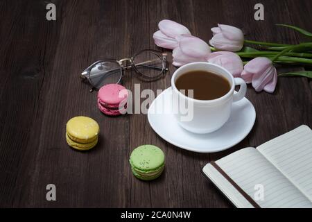 Tazza di tè, macaron, bicchieri, tulipani rosa e taccuino su sfondo di legno. Foto Stock