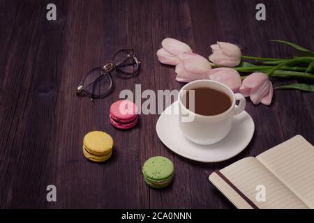 Tazza di tè, macaron, bicchieri, tulipani rosa e taccuino su sfondo di legno. Foto Stock