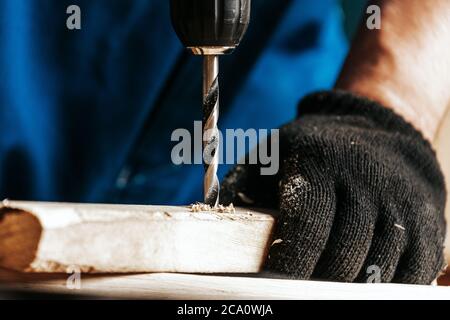 Primo piano di un operaio di costruzione maschio in una costruzione sporca nera nel complesso chiude una vite con un cacciavite moderno nero in un blocco di legno nel w Foto Stock