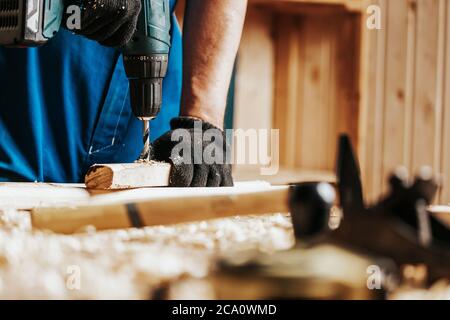 Primo piano di un operaio di costruzione maschio in una costruzione sporca nera nel complesso chiude una vite con un cacciavite moderno nero in un blocco di legno nel w Foto Stock