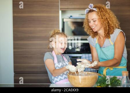 Cucina casalinga cibo. La madre allegra e la figlia di suo figlio in grembiule hanno i capelli ricci che impastano l'impasto in una grande ciotola e si divertono in cucina Foto Stock