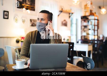 Giovane uomo con maschera facciale e computer portatile all'interno del bar, utilizzando uno smartphone. Foto Stock