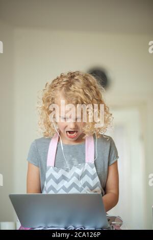 Bambina di sei anni che guarda film con sconvolgente espressione del volto stupefatto utilizzando laptop e auricolari in interni. Foto Stock