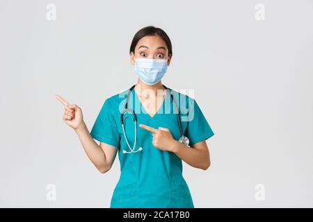 Covid-19, malattia del coronavirus, concetto degli operatori sanitari. Sorpresa e interessato asian femmina medico, medico in maschera medica e scrub puntando Foto Stock