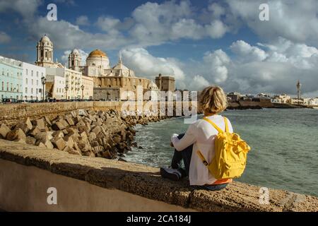 Felice viaggiatore femminile con zaino godendo di una vista sul lungomare della città vecchia e la Cattedrale di Santa Cruz in giornata di sole a Cadice, Andalusia, Spagna.Europo Foto Stock