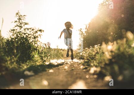 Un ragazzino corre lungo la strada al tramonto godendo della vita e della natura. Felice capretto in campagna. Silhouette di un ragazzo nei raggi solari. Aria fresca, concetto di ambiente. Foto Stock