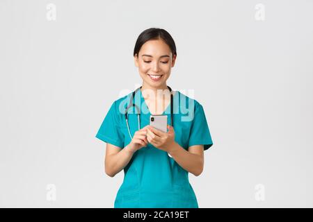 Covid-19, operatori sanitari e prevenzione virus concetto. Sorridente felice bella intern femminile asiatica, medico che fa la telefonata, guardando il cellulare Foto Stock