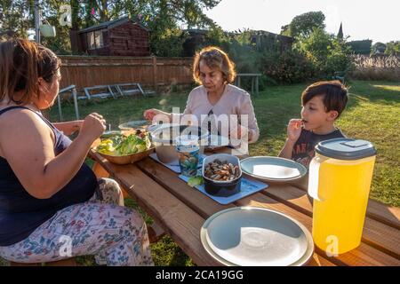 Una famiglia mangia un pasto insieme su un tavolo di trespolo fuori nel giardino posteriore. Foto Stock