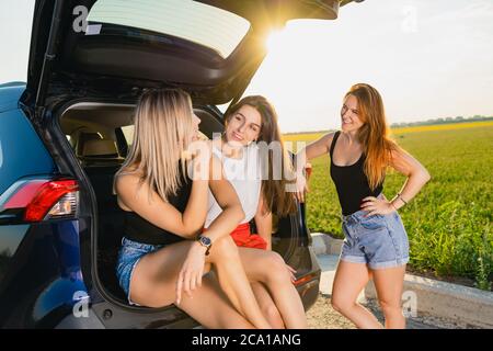 Tre belle giovani amiche si fermano durante il viaggio su strada con campo verde e tramonto, sedersi al tronco auto e parlare insieme. Foto Stock