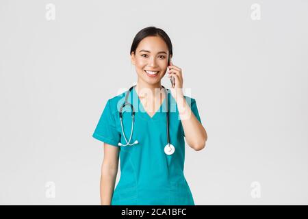 Covid-19, operatori sanitari e prevenzione virus concetto. Sorridendo felice medico asiatico, giovane intern in scrub parlando al telefono, guardando Foto Stock