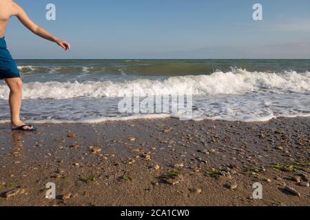 Un ragazzo in pantaloncini blu e ciabatte da spiaggia accidentalmente entrato nel telaio sulla riva in serata. Foto Stock