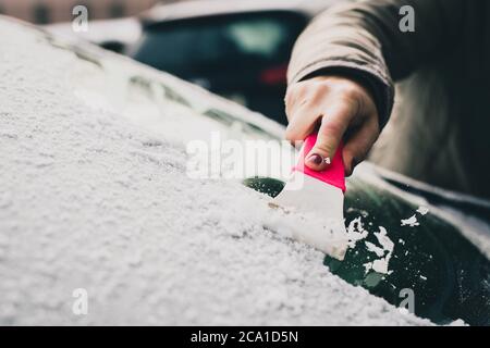 Una donna sta pulendo una finestra ghiacciata su una macchina con un raschietto di ghiaccio. Mettere a fuoco sul raschiatore di ghiaccio. Freddo nevoso e gelido mattina. Foto Stock