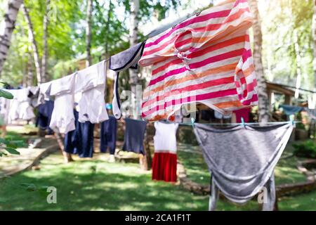 Scena di vita reale domestica di molti bambini e adulti vestiti puliti lavati appesi su stendibiancheria di betulla con spille. Cortile di casa su luminoso sole Foto Stock