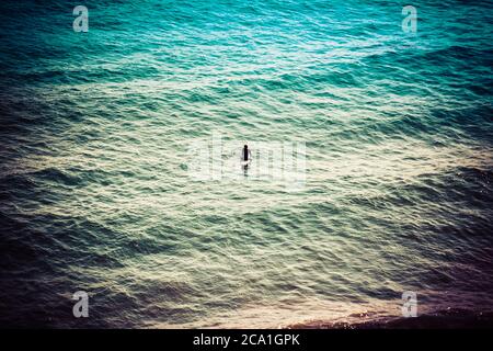 Vista rialzata e posteriore di un surfista solista in Silhouette, seduto su una tavola da surf di fronte all'orizzonte in attesa di un'onda nelle acque calme dell'Oceano Pacifico Foto Stock