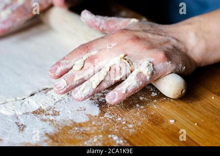 mani femminili usando un perno di laminazione per preparare un impasto su un pannello di legno sporco di farina. Preparazione di alimenti a base di farina come pane o pizza Foto Stock