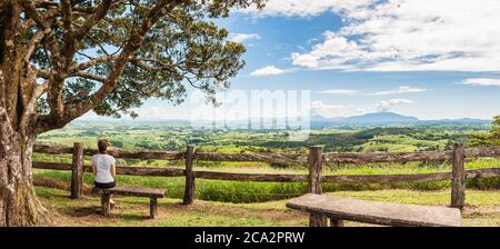 Lussureggiante paesaggio rurale panoramico con una femmina seduta su una panchina di pietra di fronte a una rustica recinzione in legno scavata a mano presso Millaa Millaa Lookout nel Queensland. Foto Stock