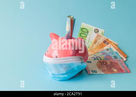 Pink piggy bank, indossando maschera protettiva, e banconote in euro isolato su sfondo blu. Banca di piggy con maschera medica usa e getta e banconote in euro. Foto Stock