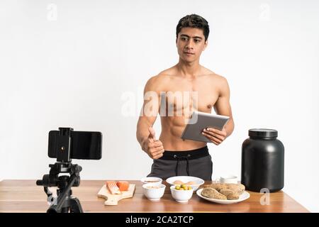 salute dell'uomo muscolare e blogger sport con pollici in su mostrando menu di cibi sani mentre si tiene un pad stand rivolto in avanti e guarda la videocamera su uno sfondo isolato Foto Stock