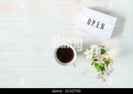 Parola APERTA su scatola luminosa, tazza di caffè e vaso con fiori su tavolo di legno. Concetto di apertura dopo la quarantena. Libertà dopo l'auto-isolamento. Se Foto Stock