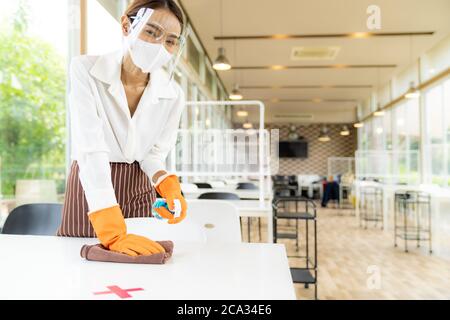 Ritratto attraente cameriera asiatica indossare maschera viso e viso schermo piano di pulizia con alcol e salvietta bagnata prima di accogliere il cliente. Nuovo normale