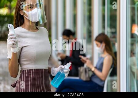 Ritratto attraente cameriera asiatica indossare maschera viso e scudo tenere termometro e igienizzatore gel di alcol con sfondo di cliente di distanza sociale