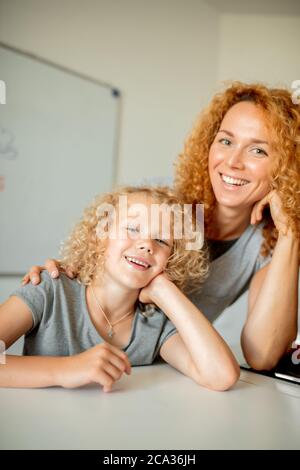 Ritratto interno di sorprendente allegra mamma caucasica con lunghi capelli ricci e la sua bambina carina molto simile a sua madre seduta al tavolo A. Foto Stock