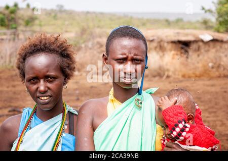 Due giovani donne Maasai, con un bambino, con un abbigliamento tradizionale, in un villaggio maasai. Riserva Nazionale di Maasai Mara. Kenya. Africa. Foto Stock