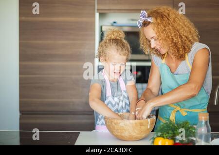 Cucina casalinga cibo. La madre allegra e la figlia di suo figlio in grembiule hanno i capelli ricci che impastano l'impasto in una grande ciotola e si divertono in cucina Foto Stock