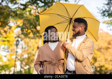 Felice coppia afro che cammina sotto ombrello nel parco autunnale Foto Stock