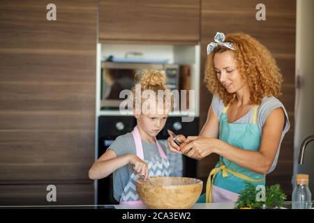 Caucasica Madre e piccola ragazza emozionale che coglie le uova alla farina per l'impasto per il compleanno del padre. Madre insegnanti sua figlia come fare in casa Foto Stock