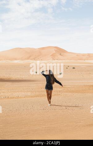 Donna che cammina nel bel deserto con dune di sabbia all'orizzonte. Viaggi in Marocco, Sahara, Merzouga. Libertà e concetto di viaggio. Foto Stock