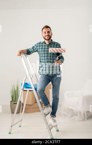 Ritratto di allegro giovane uomo bearded in piedi con rullo di vernice sulla scala in nuovo appartamento con roba in movimento Foto Stock