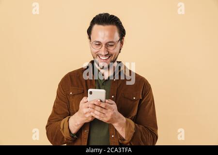 Immagine di un uomo con setole eccitate negli occhiali sorridenti e che utilizza un cellulare isolato su sfondo beige Foto Stock