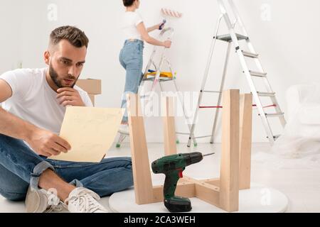 Serio giovane uomo sopportato seduto con gambe incrociate sul pavimento e lettura istruzioni durante l'assemblaggio di mobili in nuovo appartamento Foto Stock