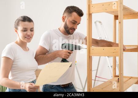 Soddisfare giovane donna che legge l'istruzione mentre assiste il marito per assemblare i mobili in nuovo appartamento Foto Stock