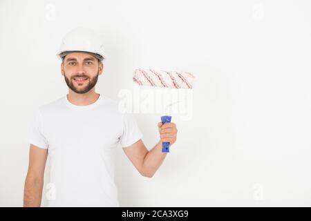 Ritratto di sorridente bel giovane decoratore maschio con stoppia indossando hardhat tenendo rullo di vernice contro parete bianca Foto Stock