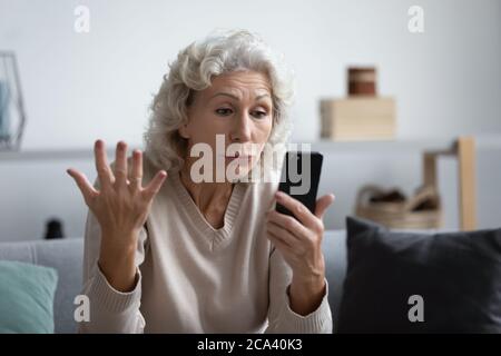 La nonna che tiene lo smartphone guarda lo schermo del dispositivo si sente arrabbiata Foto Stock