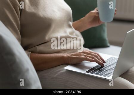 Donna anziana che riposa sul divano bevendo il tè usando il computer portatile Foto Stock