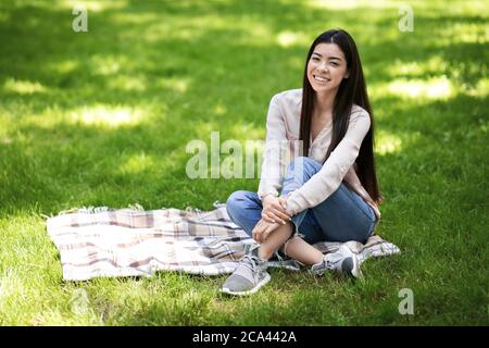 Relax all'aperto. Ritratto di giovane ragazza asiatica seduta su Plaid nel Parco Foto Stock