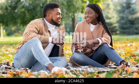 Coppia nera seduta al picnic nel parco autunnale e bere caffè Foto Stock
