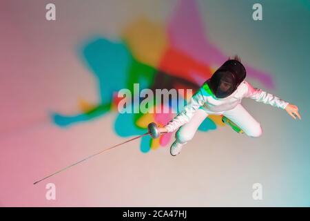 Colore. Ragazza teenager in costume da scherma con spada in mano su sfondo gradiente in luce al neon. Vista dall'alto. Giovane modello di pratica, formazione in movimento, azione. CopySpace. Sport, gioventù, stile di vita sano. Foto Stock