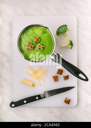 Zuppa di broccoli con crostini in pentola su tagliere bianco. Guarnice e coltello tritati. Scatto in testa. Ricetta. Foto Stock