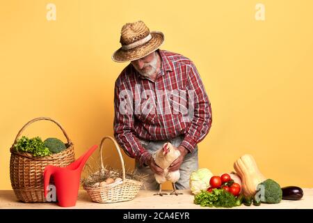 Vista frontale del contadino locale maturo, che impacchettava il suo ricco raccolto autunnale di verdure fresche e uova di organis, tenendo la sua gallina dungheill, vestito in paglia ha Foto Stock