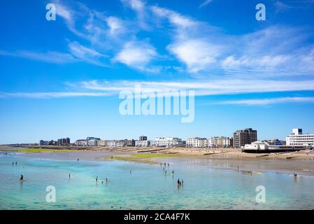 Vista costiera con bassa marea con persone che si addormellano in acque poco profonde con il cielo blu e le persone che godono l'estate sulla spiaggia durante la chiusura a Worthing, Foto Stock