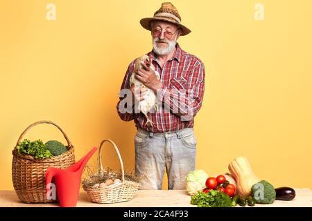 L'agricoltore maschile senior è venuto sul mercato con verdure fresche e uova dalla sua fattoria locale, tenendo con cura la sua gallina dungheill, in attesa di acquirenti, sorridendo Foto Stock