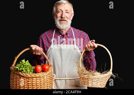 Ritratto di bel contadino maschile con rughe e barba grigia, vestito in grembiule e camicia controllata, si alza con due cesti in vimini in entrambe le mani con Foto Stock