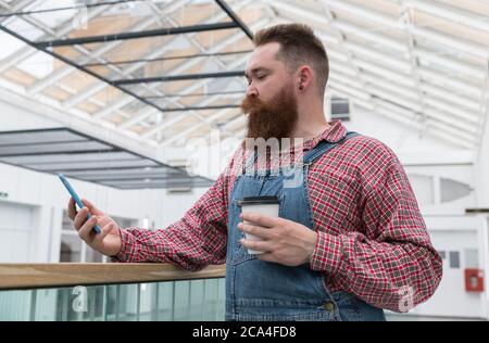 Lavoratore autonomo con cuscinetti brutali in tute blu, maglietta controllata, caffè da una tazza di carta, utilizzando smartphone. È il momento di fare una pausa. Hipster b Foto Stock